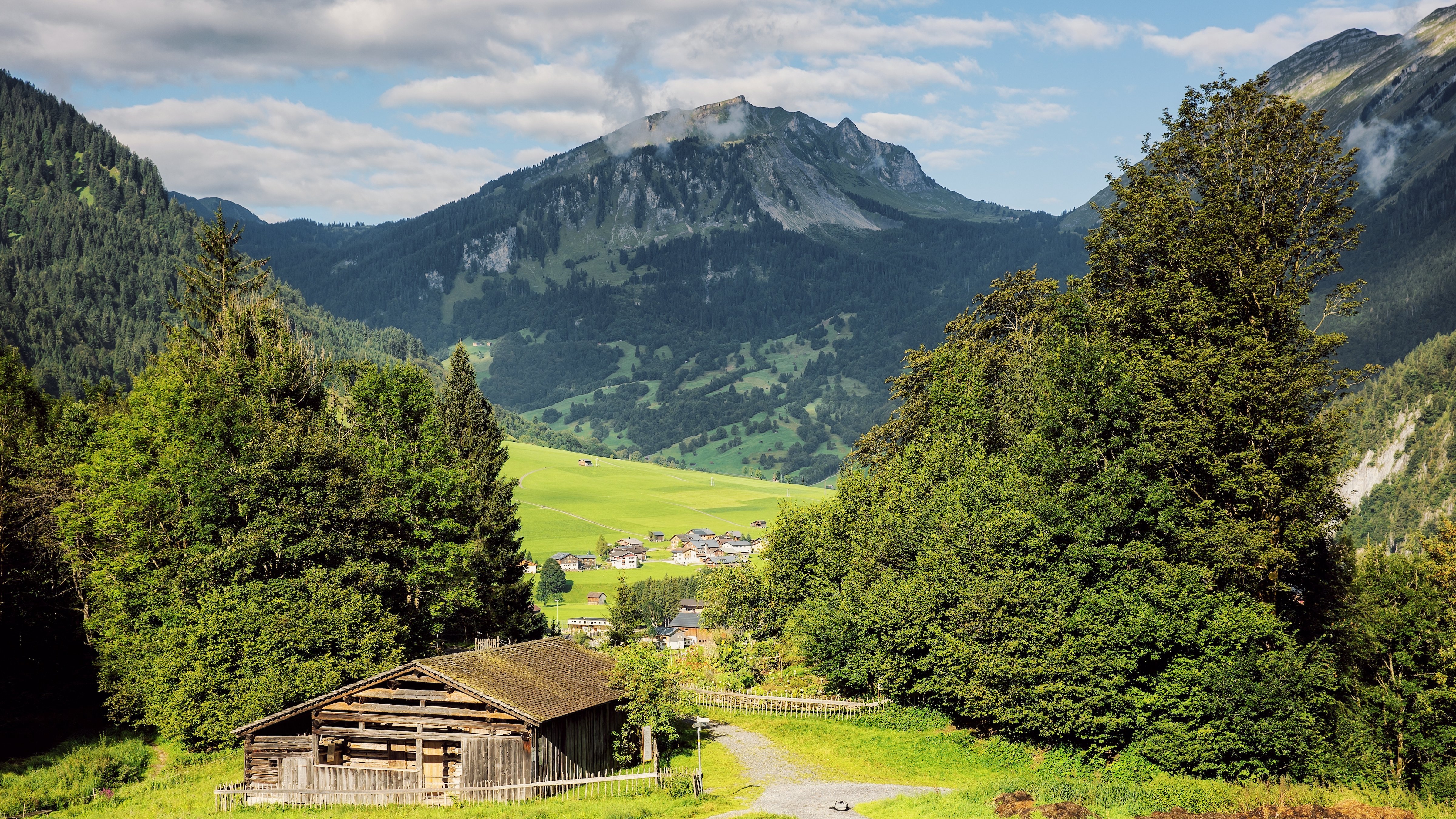 Panoramaaufnahme einer Berglandschaft mit grünen Wiesen und einem Holzgebäude im Vordergrund