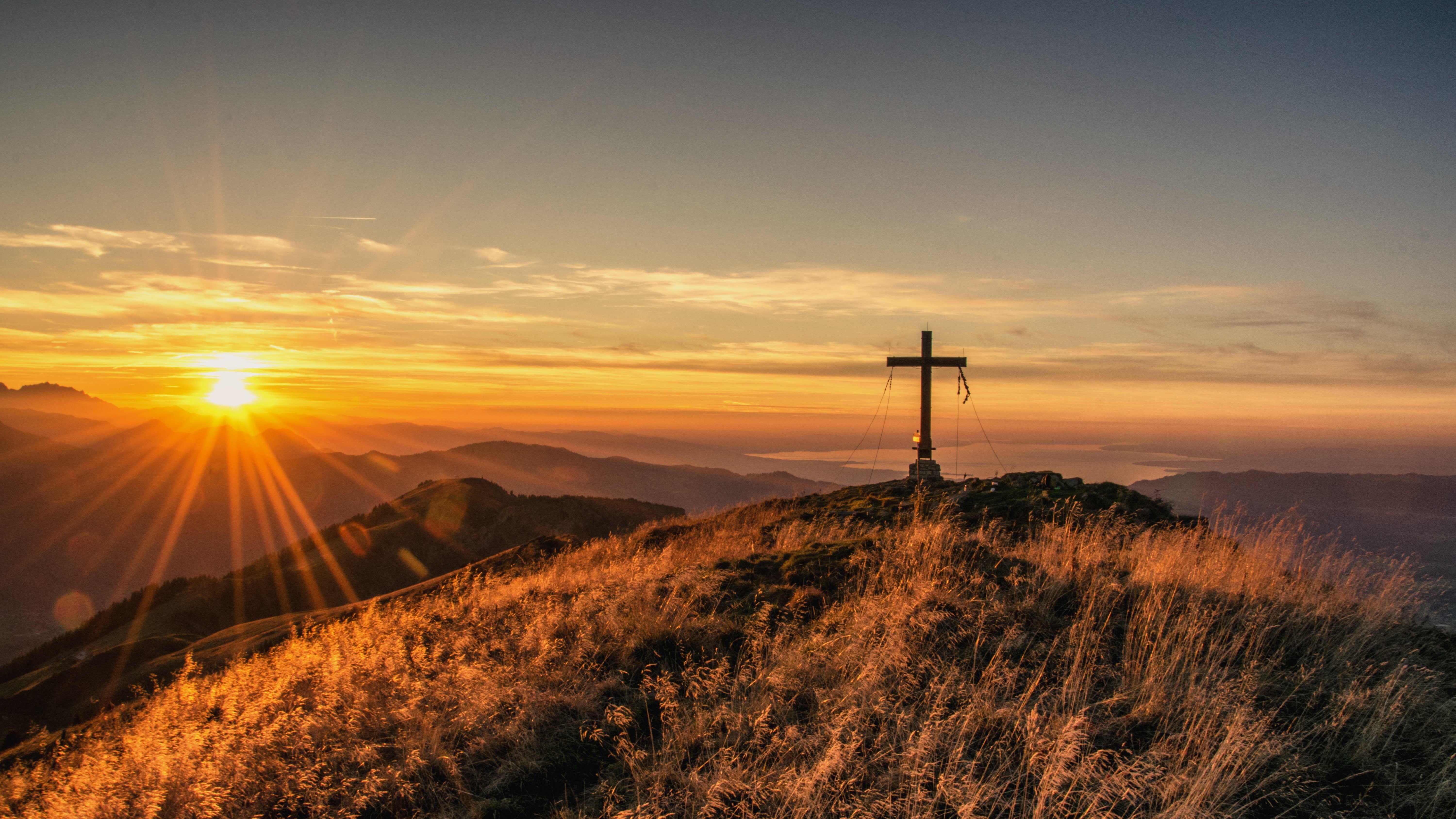 Morgendlicher Ausblick auf die Berge Ein Kreuz auf einem Hügel vor der Kulisse der Berge während des Sonnenaufgangs im Bregenzerwald.