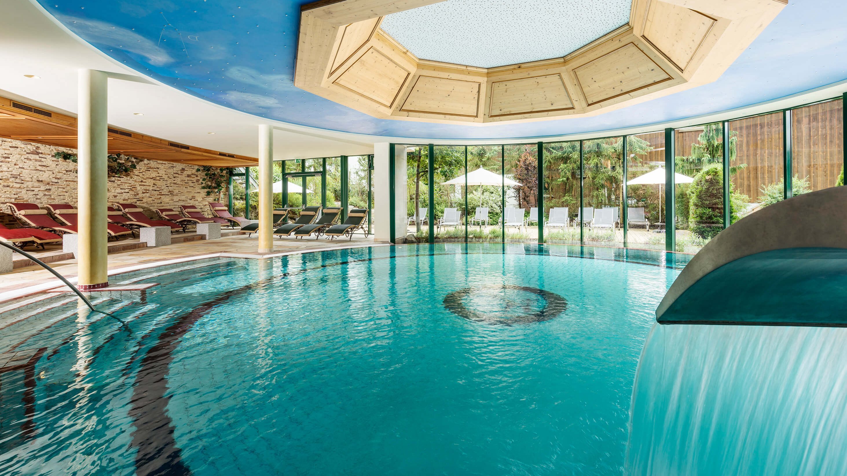 Indoor hotel spa featuring a circular pool with waterfall and lounge chairs overlooking the garden